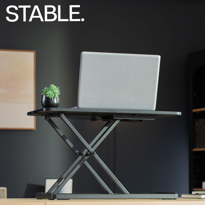 Laptop on a adjustable desk with a plant, against a dark wall